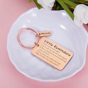 Op de afbeelding: Roze gouden sleutelhanger met een rechthoekig label waarop "Kleine herinneringen" staat en de tekst "Je bent genoeg. Het is ok&eacute; om slechte dagen te hebben. Geloof niet alles wat je denkt. Je bent zo erg geliefd. Wees vriendelijker tegen jezelf." De sleutelhanger heeft ook een kleiner label met de tekst "Wendy".