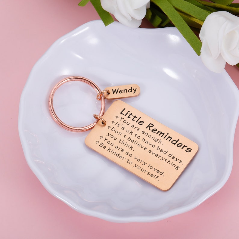 Op de afbeelding: Roze gouden sleutelhanger met een rechthoekig label waarop "Kleine herinneringen" staat en de tekst "Je bent genoeg. Het is ok&eacute; om slechte dagen te hebben. Geloof niet alles wat je denkt. Je bent zo erg geliefd. Wees vriendelijker tegen jezelf." De sleutelhanger heeft ook een kleiner label met de tekst "Wendy".