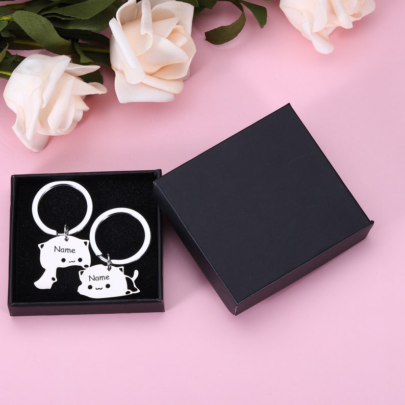 May include: Two silver cat-shaped keychains with the word "Name" engraved on each. The keychains are in a black box with a white background.