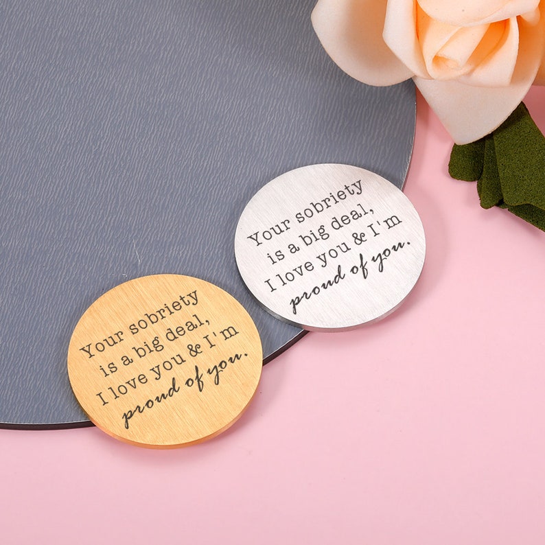 May include: Two round wooden and metal tokens with the text "Your sobriety is a big deal, I love you & I'm proud of you."  The tokens are on a pink and gray surface.