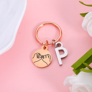 May include: A rose gold keychain with a circle charm featuring two hands making a pinky promise. The keychain also has a silver charm with the letter 'B' on it.