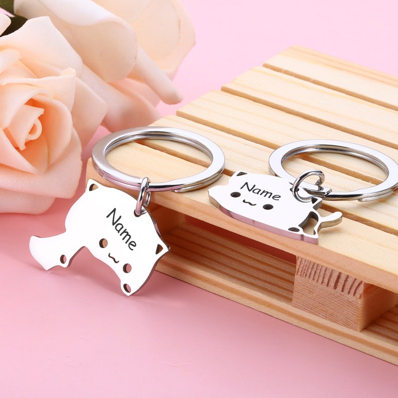 May include: Two silver keychains with cat-shaped charms. The charms have the word "Name" engraved on them. The keychains are on a wooden surface.