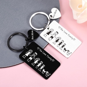 May include: Two rectangular metal keychains, one silver and one black, each featuring a split ring and a heart-shaped charm engraved with "Best Mom". The rectangular tags display a line-art family illustration and the text "The Love Family".