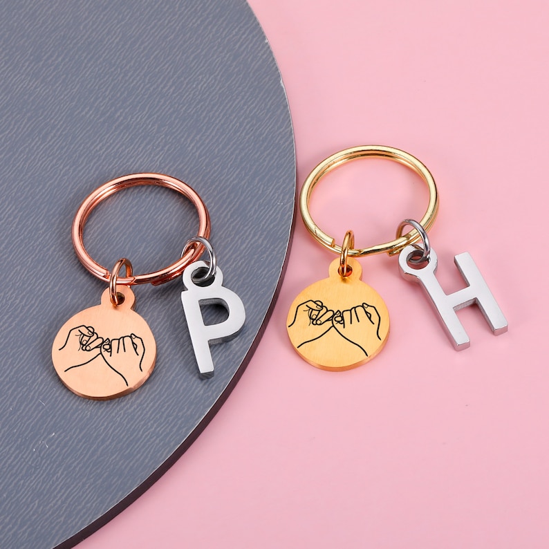 May include: Two gold keychains with a pinkie promise design and letter charms. The keychain on the left has a silver ring and a silver letter P charm. The keychain on the right has a gold ring and a silver letter H charm.