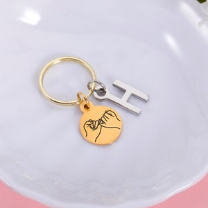 May include: Gold keychain with a small charm featuring two hands making a pinky promise. The charm is attached to a gold key ring with a silver letter H charm.