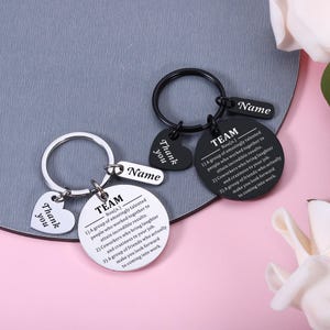 May include: Two metal keychains, one silver and one black, each featuring a round disc engraved with the definition of TEAM, a heart-shaped charm engraved with Thank you, and a small rectangular tag with the word Name engraved on the surface.