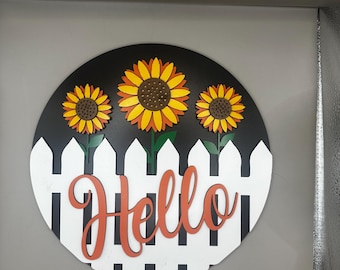 May include: A round wooden sign with a black and white picket fence design and the word "Hello" in orange. Three yellow sunflowers with orange centers are placed on top of the fence.