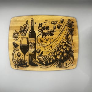 May include: Wooden cutting board with a laser-etched design featuring a wine bottle, a wine glass, grapes, and the words "Bon Appetit." The board has a rectangular shape with rounded edges, perfect for serving appetizers.