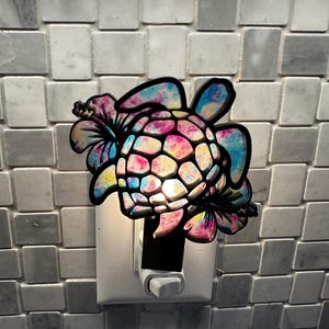 Op de afbeelding: Een kleurrijk tie-dye zeeschildpad nachtlampje met een hibiscusbloem ontwerp. Het nachtlampje is aangesloten op een stopcontact.