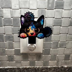 May include: A colorful nightlight shaped like a cat peeking out from behind a light switch. The cat has black fur with blue, purple, and yellow accents. The nightlight has a flower on its head.