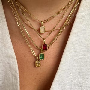 May include: Four gold necklaces with different coloured gemstone pendants. The pendants are all rectangular in shape. The necklaces are layered on top of each other. The top pendant is white, the second is red, the third is green, and the bottom is yellow.