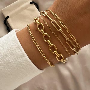May include: Several gold bracelets of different chain designs, such as a fine chain, a curb chain, and a paperclip chain. The bracelets are worn on a wrist, with a white shirt and a small white bag visible in the background.