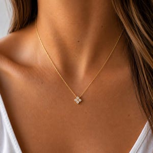 May include: A gold necklace with a four-petal flower pendant set with small gemstones. The necklace is worn on a person's chest, with the chain against the skin. The pendant is central, and the gold chain is fine and elegant.