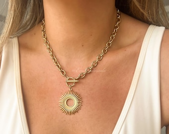 May include: Gold-toned necklace featuring a large sunburst pendant and a chunky chain. The pendant has a circular center with radiating spikes. The necklace is secured with a toggle clasp. The jewelry is worn against a white top.