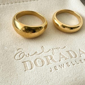 Könnte beinhalten: Zwei einfache, abgerundete goldfarbene Eheringe auf einer weißen, strukturierten Oberfläche. Die Oberfläche trägt den Text "Evelyn Dorada Jewellers" in Prägung.