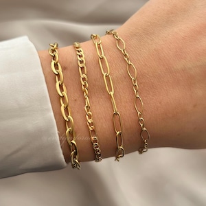 May include: Four gold chain bracelets on a person's wrist. The bracelets are different styles and thicknesses.
