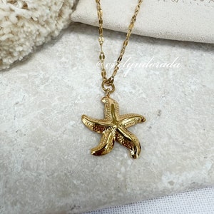 Könnte beinhalten: Goldkette mit einem goldenen Seestern-Anhänger. Die Kette liegt auf einer weißen Oberfläche mit einer kleinen weißen Tasche, auf der "Evelyn Dorada Jewelry" steht.