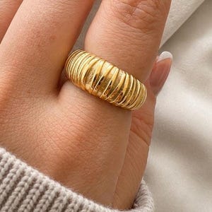 May include: A gold-toned ring featuring a wide, domed band with a series of vertical, parallel ridges or grooves that wrap around the entire circumference of the piece, creating a textured, ribbed appearance with a polished, reflective finish.