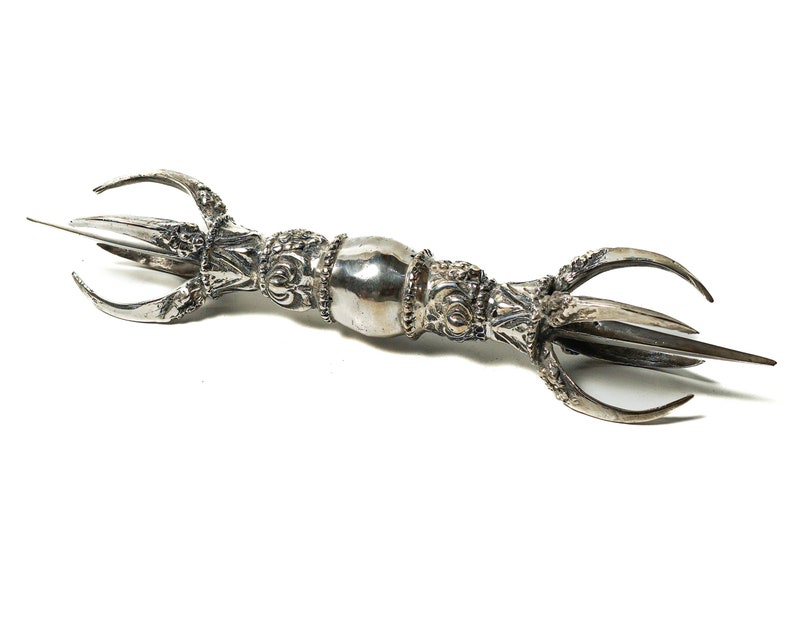 May include: A silver-colored ceremonial weapon with a three-pronged head and intricate details. The weapon is known as a "vajra" and is a symbol of power and protection in Buddhism.