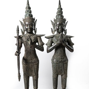 Op de afbeelding: Twee donkerbronzen beelden van figuren in traditionele Thaise kleding, staand met gevouwen handen in gebed. De figuren zijn versierd met ingewikkelde details en dragen uitgebreide hoofdtooien.