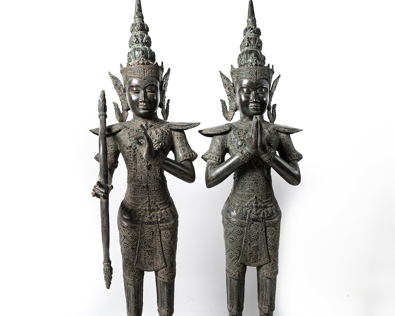 Op de afbeelding: Twee donkerbronzen beelden van figuren in traditionele Thaise kleding, staand met gevouwen handen in gebed. De figuren zijn versierd met ingewikkelde details en dragen uitgebreide hoofdtooien.