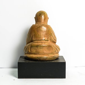 May include: A small, ceramic statue of a seated figure with a brown and green glaze. The figure is wearing a robe and has a serene expression. The statue is sitting on a black wooden base.