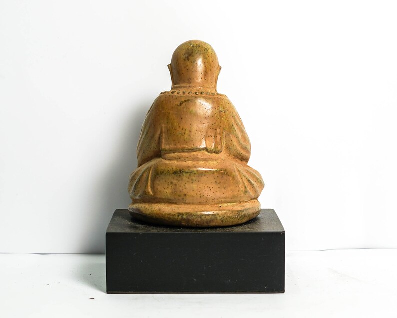 May include: A small, ceramic statue of a seated figure with a brown and green glaze. The figure is wearing a robe and has a serene expression. The statue is sitting on a black wooden base.