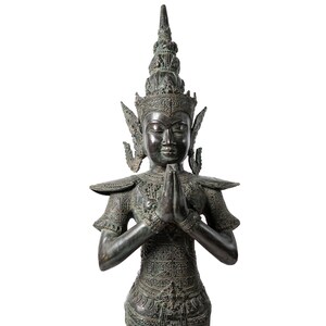 Op de afbeelding: Een donker bronzen beeld van een godheid met een hoge, sierlijke kroon en handen die in gebed zijn gevouwen. Het beeld is gedetailleerd met ingewikkelde houtsnijwerk en heeft een serene uitdrukking.