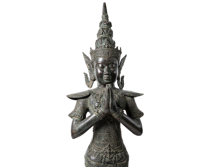 Op de afbeelding: Een donker bronzen beeld van een godheid met een hoge, sierlijke kroon en handen die in gebed zijn gevouwen. Het beeld is gedetailleerd met ingewikkelde houtsnijwerk en heeft een serene uitdrukking.