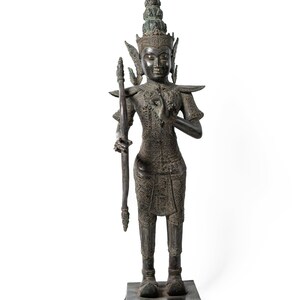 Op de afbeelding: Een donker bronzen beeld van een staande figuur met een kroon en een boog en pijl. De figuur draagt ​​uitgebreide kleding en sieraden. Het beeld staat op een vierkante basis.