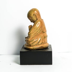 May include: A small, ceramic statue of a person in a seated position with their hands clasped together. The statue is painted in shades of brown and green, and is sitting on a black base.