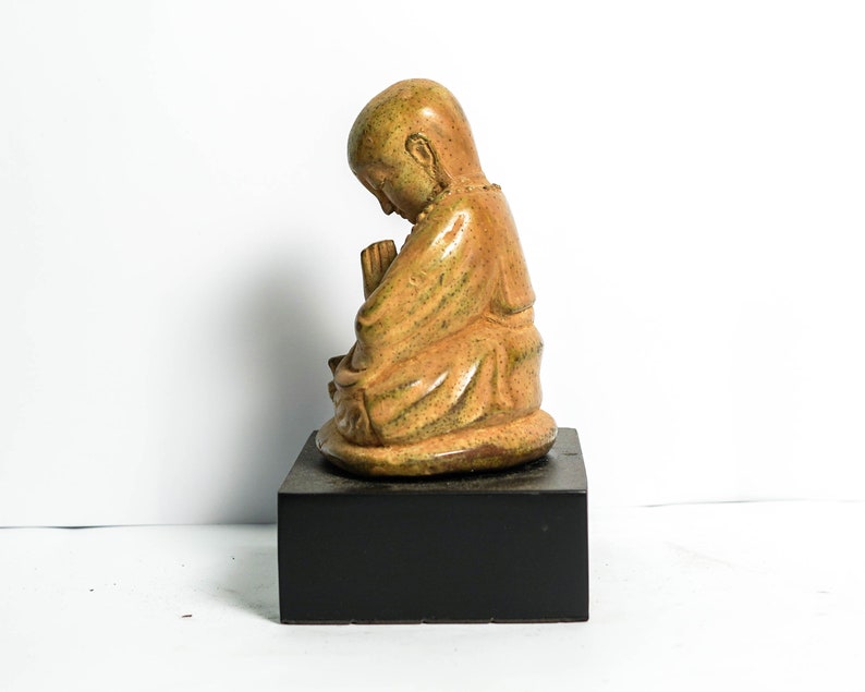 May include: A small, ceramic statue of a person in a seated position with their hands clasped together. The statue is painted in shades of brown and green, and is sitting on a black base.