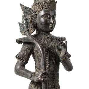 Op de afbeelding: Een gedetailleerd bronzen beeld van een godheid met een kroon en ingewikkelde houtsnijwerk. Het beeld houdt een staf vast en heeft een serene uitdrukking.