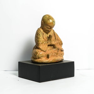 May include: A small, seated, golden-brown ceramic statue of a monk with hands clasped in prayer. The statue is on a black wooden base.
