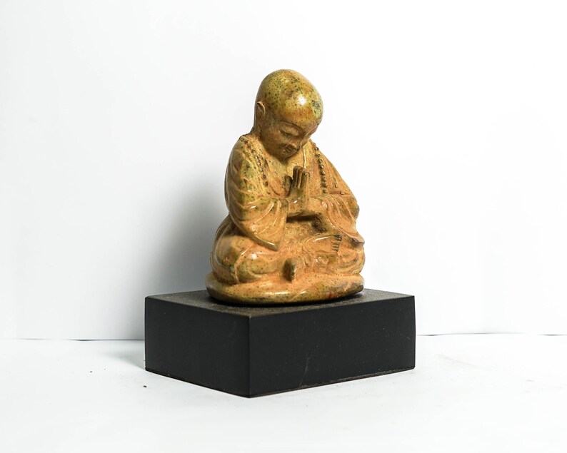 May include: A small, seated, golden-brown ceramic statue of a monk with hands clasped in prayer. The statue is on a black wooden base.