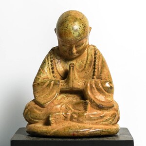 May include: A small, ceramic statue of a seated Buddha with a speckled, yellow-brown glaze. The Buddha is in a meditative pose with hands clasped in front of the chest.