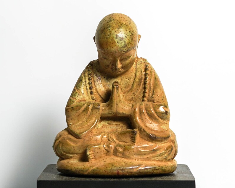 May include: A small, ceramic statue of a seated Buddha with a speckled, yellow-brown glaze. The Buddha is in a meditative pose with hands clasped in front of the chest.