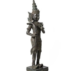 Op de afbeelding: Een donker bronzen beeld van een staande figuur met een kroon en een uitgebreide hoofdtooi. De figuur houdt een boog en pijl vast en heeft een gedetailleerd, versierd ontwerp.