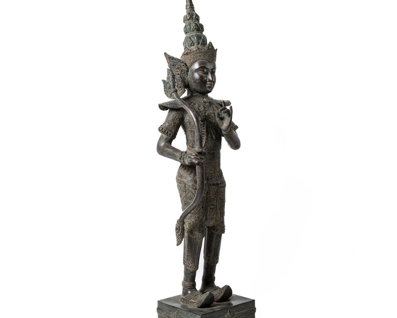 Op de afbeelding: Een donker bronzen beeld van een staande figuur met een kroon en een uitgebreide hoofdtooi. De figuur houdt een boog en pijl vast en heeft een gedetailleerd, versierd ontwerp.