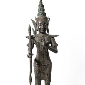 Op de afbeelding: Een donker bronzen beeld van een godheid met een kroon en uitgebreide sieraden, die een boog en pijl in een gebedshouding vasthoudt.