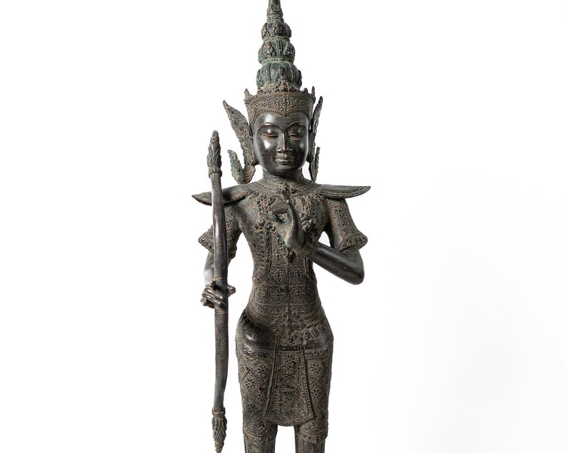 Op de afbeelding: Een donker bronzen beeld van een godheid met een kroon en uitgebreide sieraden, die een boog en pijl in een gebedshouding vasthoudt.