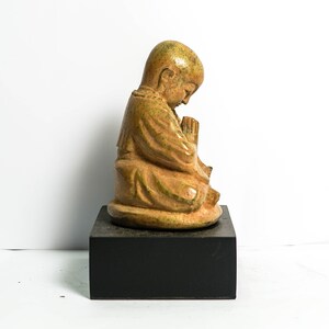 May include: A small, ceramic statue of a monk in a seated position with hands clasped in prayer. The statue is painted in shades of brown and green and is placed on a black wooden base.