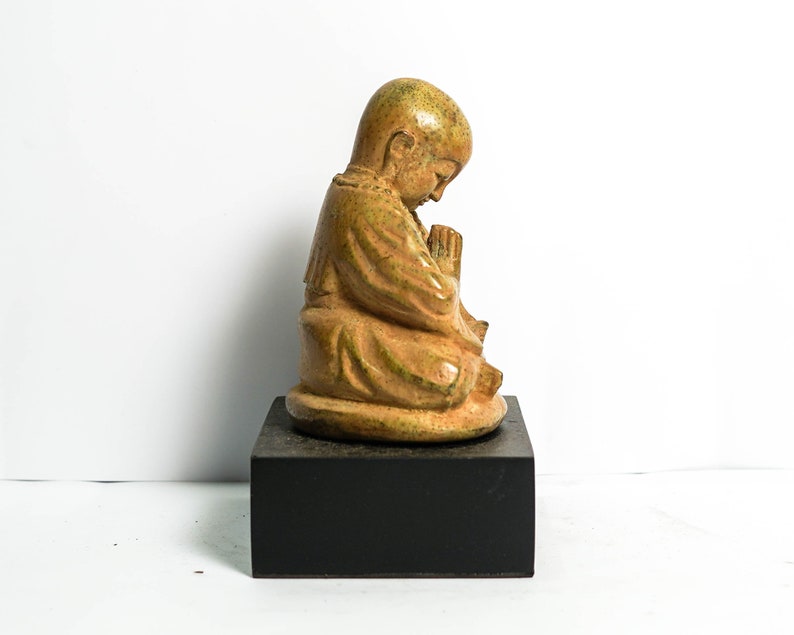 May include: A small, ceramic statue of a monk in a seated position with hands clasped in prayer. The statue is painted in shades of brown and green and is placed on a black wooden base.