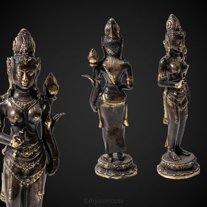 May include: Three bronze statues of a female deity with gold accents. The statues are standing on a base and have a lotus flower in their hand. The statues are in a dark background.