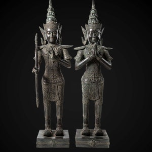 Op de afbeelding: Twee donkerbronzen beelden van figuren met uitgebreide hoofdtooien en sierlijke kleding. De figuren staan op vierkante bases en hebben hun handen gevouwen in een gebedspositie. Het beeld links houdt een speer in zijn linkerhand.