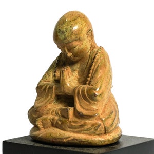 May include: A small, green and brown ceramic statue of a meditating figure with hands clasped in prayer. The figure is sitting with legs crossed and wearing a robe.