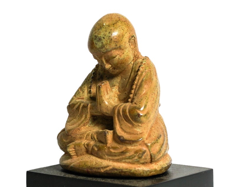 May include: A small, green and brown ceramic statue of a meditating figure with hands clasped in prayer. The figure is sitting with legs crossed and wearing a robe.