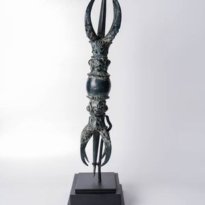 May include: A dark green metal sculpture of a stylized spear with a pointed tip and three curved prongs at the base. The sculpture is mounted on a black wooden base.