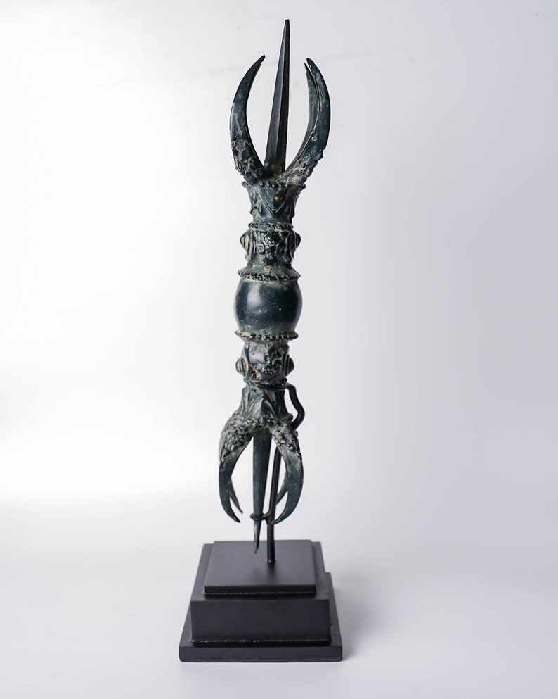May include: A dark green metal sculpture of a stylized spear with a pointed tip and three curved prongs at the base. The sculpture is mounted on a black wooden base.