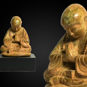 May include: A small, golden-brown ceramic statue of a meditating monk with hands clasped in prayer. The statue has a textured surface and is sitting on a black base.